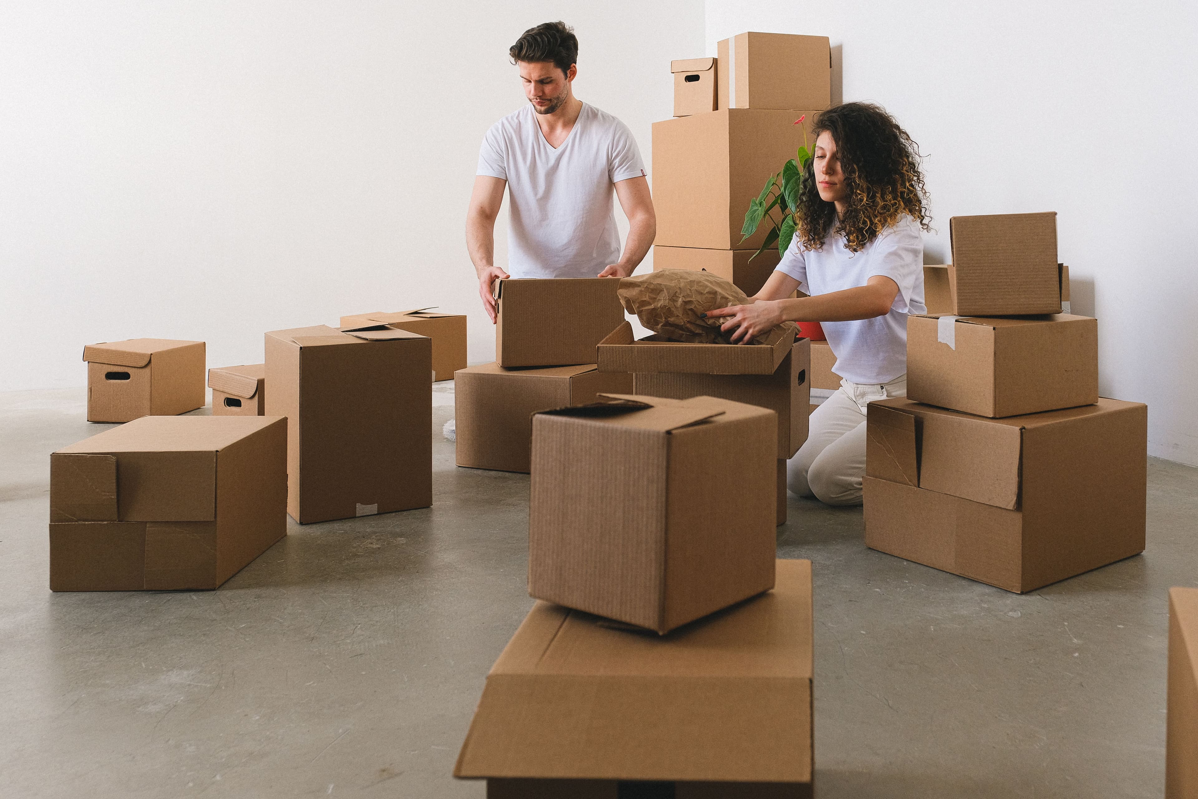 A couple packs household belongings into moving boxes in a bright apartment.